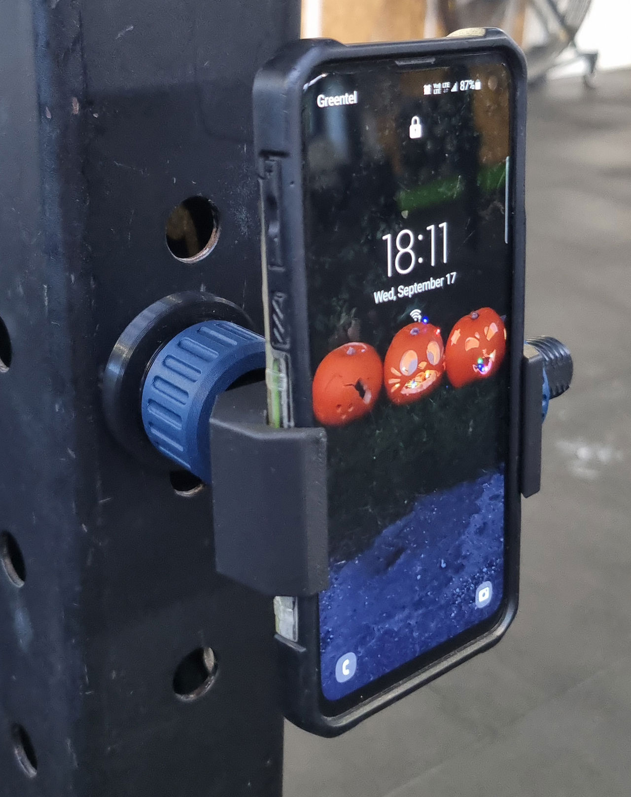 Sort smartphoneholder med blå drejeknap monteret på fitnessstativ.